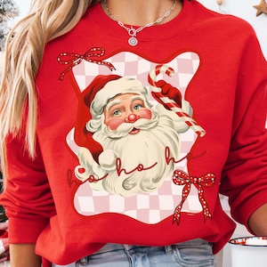 May include: Red sweatshirt featuring a Santa Claus graphic holding a candy cane. The design includes the words "ho ho ho" and red bows. The Santa illustration is set against a pink and white checkered background.