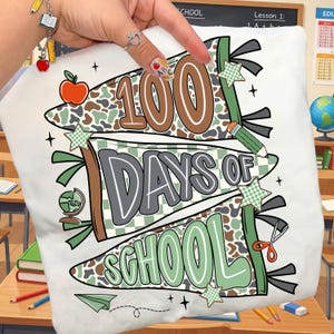 May include: White t-shirt with a colorful graphic celebrating "100 Days of School." The design features banners with the text, along with an apple, pencil, globe, scissors, and stars. The color palette includes brown, green, and gray.