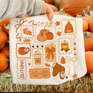 May include: A white autumn-themed decorative pillow featuring various orange and brown illustrations. The pillow includes images of pumpkins, a slice of pie, a mug, a candle, and the word "autumn". The word "cozy" is also written on the pillow.
