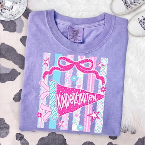 May include: A lilac Comfort Colors t-shirt featuring a colorful graphic. The graphic includes a pink pennant that reads "Kindergarten", a pink bow, stars, and striped and floral patterns. The shirt is a small size.