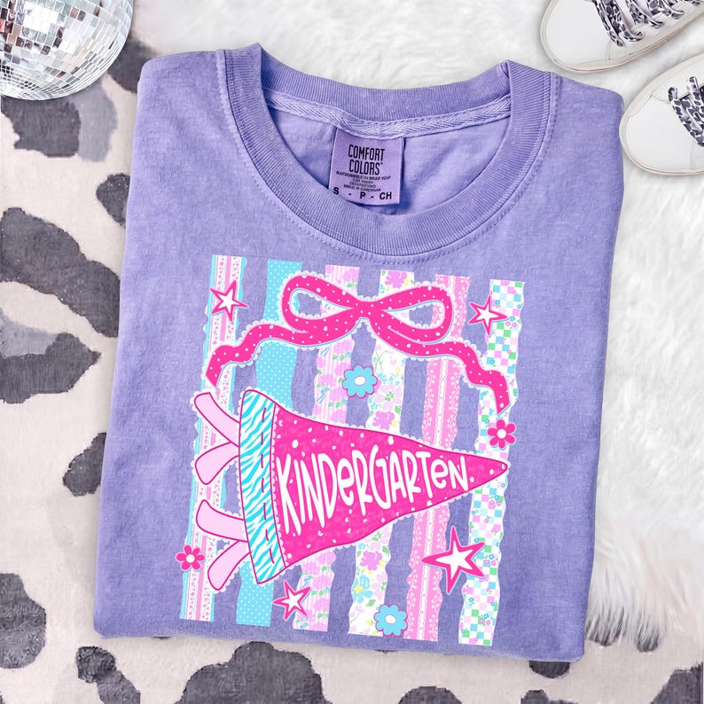 May include: A lilac Comfort Colors t-shirt featuring a colorful graphic. The graphic includes a pink pennant that reads "Kindergarten", a pink bow, stars, and striped and floral patterns. The shirt is a small size.