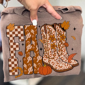 Puede incluir: Una sudadera marrón con la palabra "FALL" en un diseño con un patrón de cuadros y cactus. El diseño incluye una herradura, una calabaza y botas de vaquero con detalles florales y de leopardo. Ropa con temática otoñal.