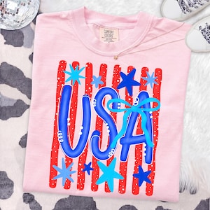 May include: A light pink t-shirt with a patriotic design. The design features the word "USA" in blue, with red and white stripes and blue stars. The shirt is made of a soft material and is perfect for celebrating the 4th of July.