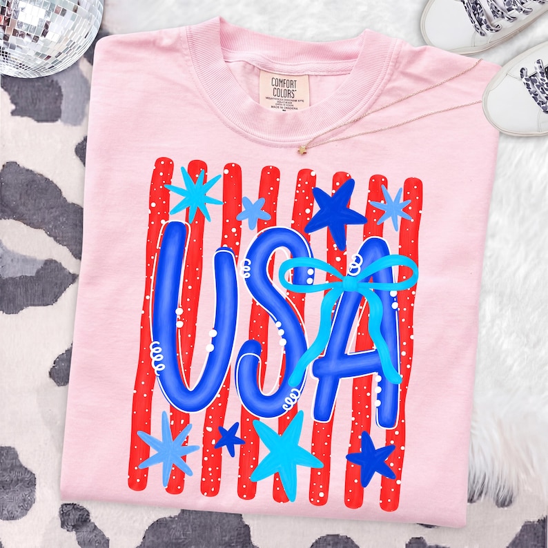 May include: A light pink t-shirt with a patriotic design. The design features the word "USA" in blue, with red and white stripes and blue stars. The shirt is made of a soft material and is perfect for celebrating the 4th of July.