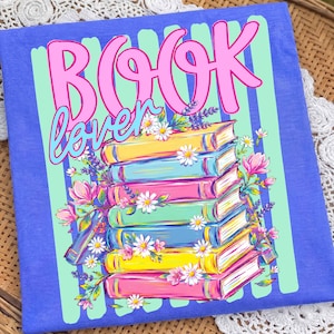 May include: A blue t-shirt featuring a graphic print of a stack of colorful books decorated with pink and white flowers. The text "BOOK lover" is printed in large pink and blue script letters above the books, set against a light green striped background.