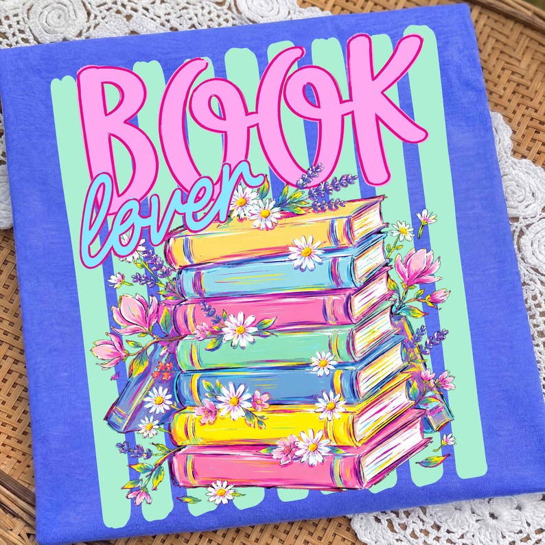 Puede incluir: Una camiseta azul con un estampado gr&aacute;fico de una pila de libros coloridos decorados con flores rosas y blancas. El texto "BOOK lover" est&aacute; impreso en grandes letras cursivas rosas y azules sobre los libros, sobre un fondo de rayas verde claro.