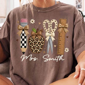 May include: A brown t-shirt featuring a graphic print of a pencil with a checkered pattern, a leopard print apple, scissors with a polka dot pattern, and a wooden ruler. Each item is adorned with a gold bow. The text "Mrs. Smith" is printed below.