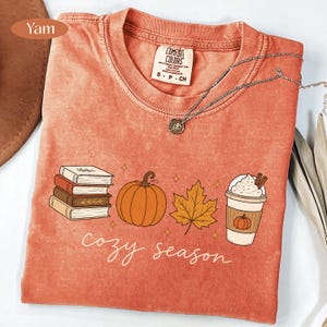 May include: A yam-colored t-shirt featuring a graphic print of a stack of books, a pumpkin, a maple leaf, and a coffee cup with whipped cream. The text "cozy season" is printed below the graphics in a white script font. A silver necklace is visible.