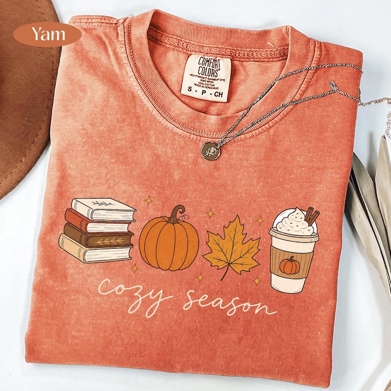 May include: A yam-colored t-shirt featuring a graphic print of a stack of books, a pumpkin, a maple leaf, and a coffee cup with whipped cream. The text "cozy season" is printed below the graphics in a white script font. A silver necklace is visible.