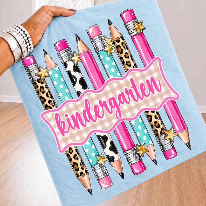 Peut inclure: Un tissu bleu clair avec un motif r&eacute;p&eacute;titif de crayons &agrave; gommes roses, des &eacute;toiles dor&eacute;es et divers designs incluant des imprim&eacute;s l&eacute;opard, vache et des pois turquoise. Une banni&egrave;re centrale &agrave; carreaux beige affiche le mot kindergarten.