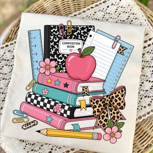 May include: A white t-shirt featuring a graphic print of a stack of books, a composition book, a ruler, a pencil, paper, paperclips, a leopard print bow, a red apple, and pink flowers. The composition book has the text COMPOSITION BOOK.