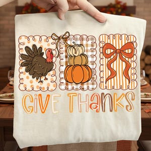 May include: A beige tote bag featuring a Thanksgiving design. The design includes a turkey, stacked pumpkins, and a bow, all within decorative frames. The words "GIVE THANKS" are printed in orange and brown.