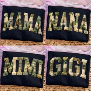 Op de afbeelding: Vier zwarte sweatshirts met verschillende namen in een camouflagepatroon. De namen zijn "MAMA", "NANA", "MIMI" en "GIGI". De sweatshirts worden gepresenteerd op een geweven oppervlak.