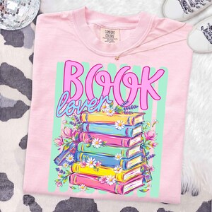 Puede incluir: Una camiseta rosa claro con un estampado gr&aacute;fico que muestra el texto "BOOK lover" en letra cursiva rosa y azul. Debajo del texto hay una pila de seis libros coloridos en amarillo, azul y rosa, decorados con margaritas blancas y lavanda.