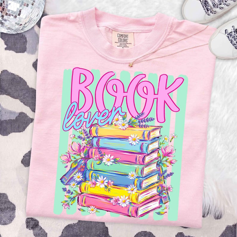 Puede incluir: Una camiseta rosa claro con un estampado gr&aacute;fico que muestra el texto "BOOK lover" en letra cursiva rosa y azul. Debajo del texto hay una pila de seis libros coloridos en amarillo, azul y rosa, decorados con margaritas blancas y lavanda.