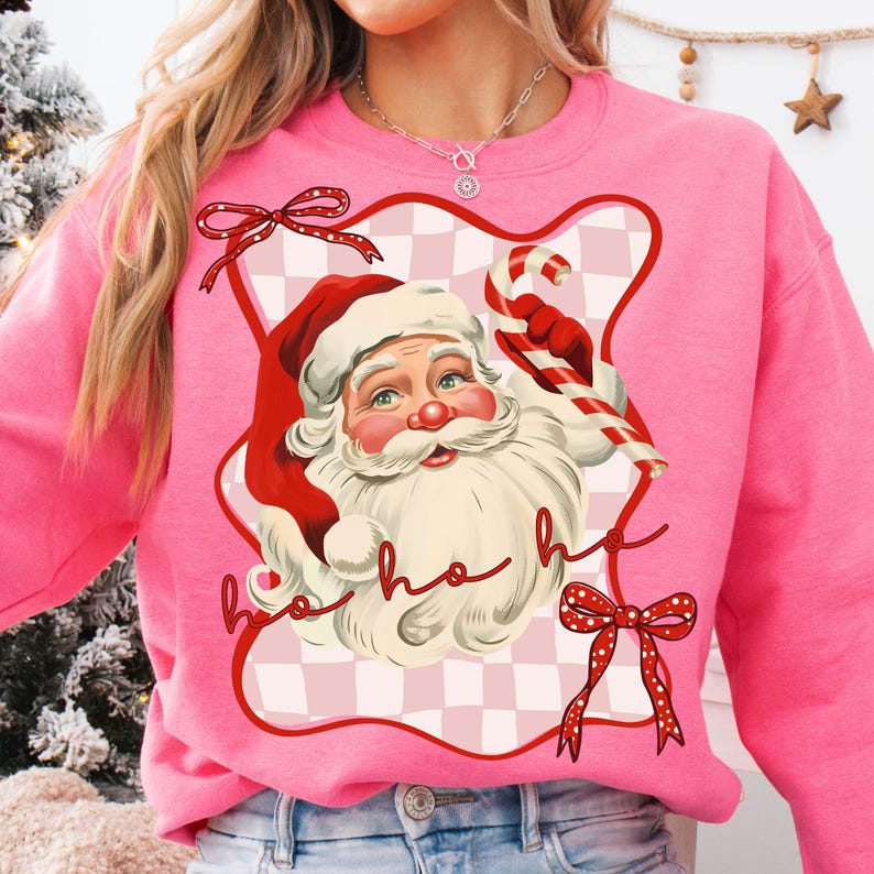 May include: Pink sweatshirt featuring a vintage Santa Claus graphic. Santa is wearing a red hat and holding a candy cane. The design includes red bows and the words "ho ho ho". The background is a pink and white checkered pattern.