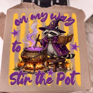 May include: A tan t-shirt featuring a graphic of a raccoon wearing a purple wizard hat and robe, stirring a bubbling cauldron with a wand while holding a book. The text reads "on my way to stir the pot" with yellow stripes, stars, and pumpkins.