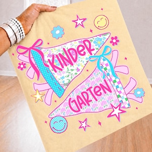 Peut inclure: Un article en tissu beige orn&eacute; de deux graphismes en forme de fanion avec le texte "KINDER" et "GARTEN" en &eacute;criture rose. Le motif comprend des fleurs bleues et roses, des carreaux, des rubans roses, des &eacute;toiles jaunes et un smiley jaune.