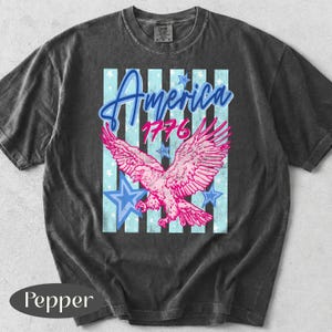 Puede incluir: Camiseta gris oscuro con un &aacute;guila rosa y el texto "America 1776". El dise&ntilde;o incluye estrellas azules y rayas verticales. La camiseta tiene un aspecto vintage y desgastado.
