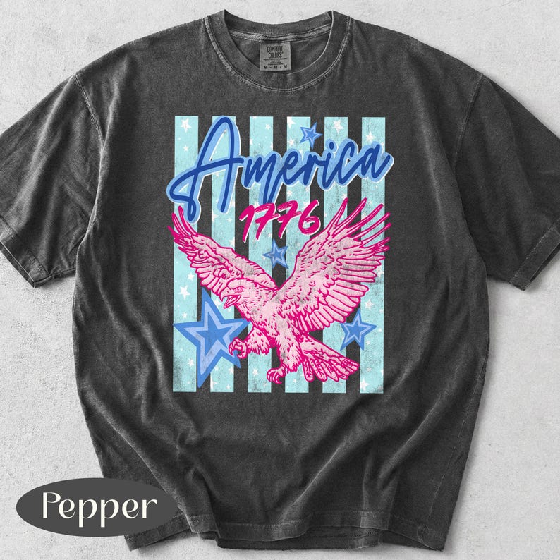 Puede incluir: Camiseta gris oscuro con un &aacute;guila rosa y el texto "America 1776". El dise&ntilde;o incluye estrellas azules y rayas verticales. La camiseta tiene un aspecto vintage y desgastado.
