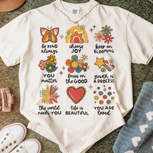 May include: White t-shirt with a colorful graphic design featuring flowers, butterflies, and positive messages like "be kind always", "choose joy", "keep on blooming", "you matter", "focus on the good", "growth is a process", "the world needs you", "life is beautiful", and "you are loved".