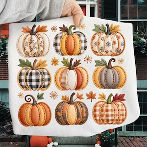 May include: White decorative kitchen towel featuring nine colorful pumpkins with various patterns and textures. The pumpkins are in shades of orange, brown, and cream, with green leaves and stems. The towel is held up by a person's hand.