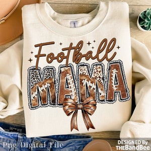 May include: A beige sweatshirt with the text "Football Mama" in brown letters with a brown and white leopard print design. The letters are decorated with brown footballs and a brown bow.