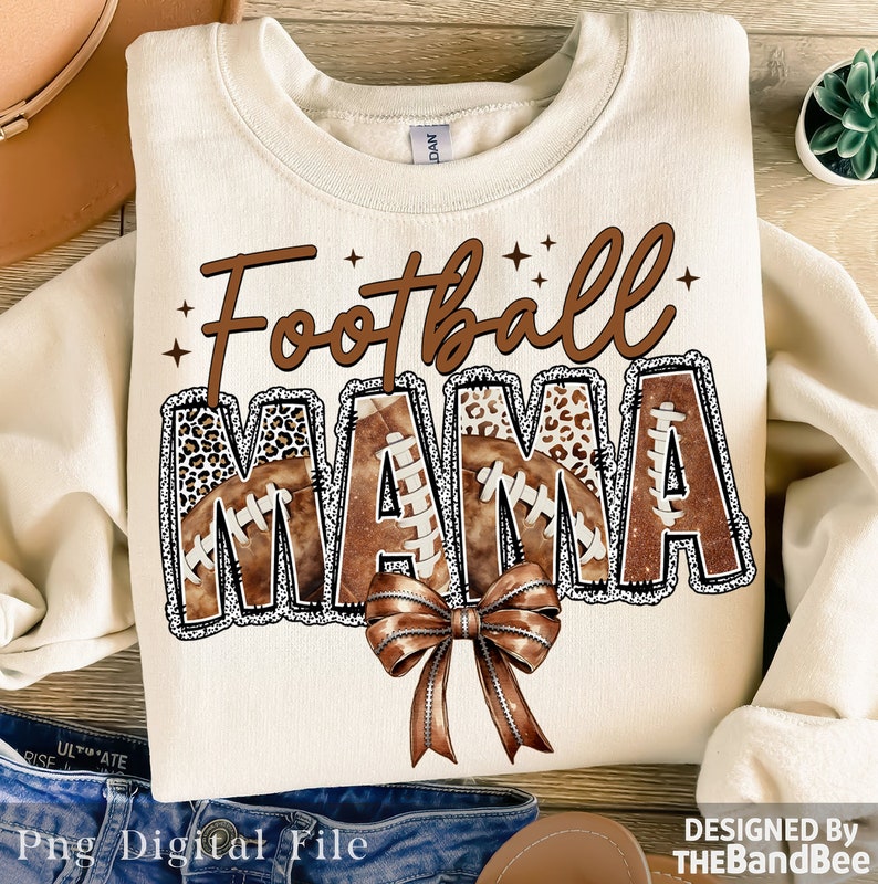 May include: A beige sweatshirt with the text "Football Mama" in brown letters with a brown and white leopard print design. The letters are decorated with brown footballs and a brown bow.