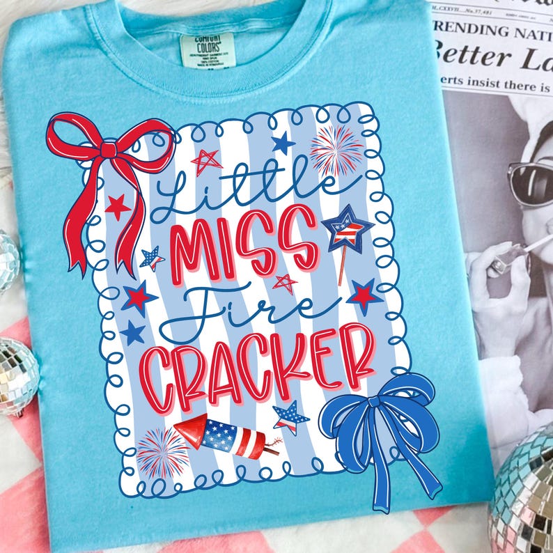 May include: Pale blue t-shirt featuring a festive design. The text "Little Miss Fire Cracker" is displayed in red and blue, accompanied by stars, fireworks, and bows. This comfortable tee is ideal for celebrating special occasions.