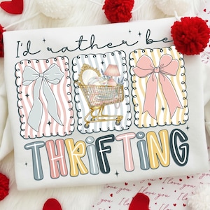 May include: White sweatshirt with a colorful graphic that says "I'd rather be THRIFTING." The graphic includes three framed images of a bow, a shopping cart filled with items, and another bow. The text is in a playful font.