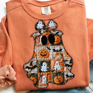 May include: Orange Comfort Colors sweatshirt with a patchwork ghost design. The ghost features pumpkins, bats, and ghost figures. The design is stitched with black thread, creating a unique Halloween-themed apparel.