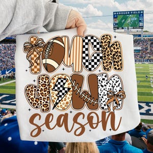 May include: White fabric with the words "TOUCHDOWN SEASON" in brown, black, and leopard print lettering. The design includes a football, lightning bolt, and bows. The background shows a football stadium.