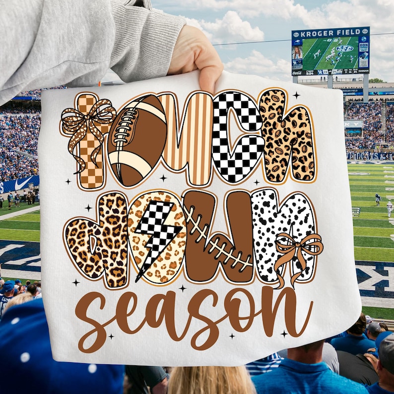 May include: White fabric with the words "TOUCHDOWN SEASON" in brown, black, and leopard print lettering. The design includes a football, lightning bolt, and bows. The background shows a football stadium.