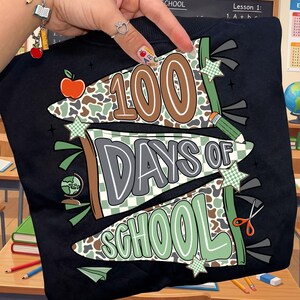 May include: Black sweatshirt with a colorful graphic celebrating 100 days of school. The design includes the text "100 Days of School" in brown, gray, and green, along with school-themed illustrations like an apple, pencil, globe, and scissors.