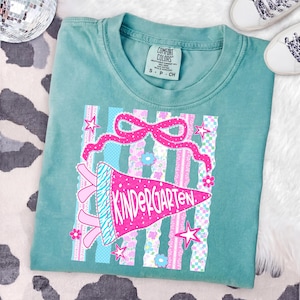 May include: Teal Comfort Colors t-shirt with a pink and blue Kindergarten pennant design. The design includes a pink bow, stars, and floral accents. The shirt is a small size.