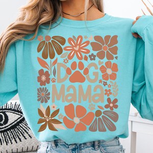 May include: A turquoise sweatshirt featuring a floral design and the text "Dog Mama". The design incorporates brown and orange flowers and paw prints. A comfortable and stylish garment.