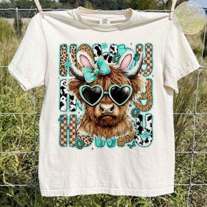 May include: Cream-colored t-shirt featuring a Highland cow graphic wearing heart-shaped sunglasses, bunny ears, and a turquoise bow. The text "Howdy Howdy Howdy" is displayed in a playful font with leopard and cow print accents.