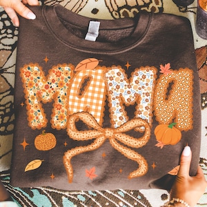 May include: A brown sweatshirt with the word "MAMA" in a decorative font. The letters are adorned with floral, gingham, and leopard print patterns. A large bow and pumpkins are also featured in the design, with fall leaves.