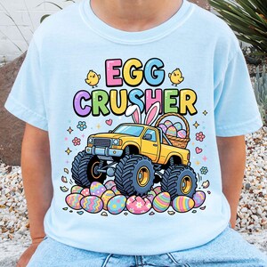 May include: Pale blue t-shirt with a colourful graphic. It shows a yellow monster truck with Easter eggs and the text "EGG CRUSHER". The truck has bunny ears and is surrounded by decorated eggs and flowers.