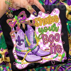 May include: Black t-shirt with a purple and white checkered border featuring a pair of purple cowboy boots with glitter accents, a fleur-de-lis, and the text "Shake your BOO-tie" in yellow and green. The boots are adorned with green and yellow tassels and beaded necklaces.