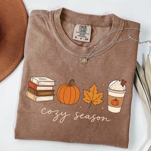 May include: A brown short-sleeved t-shirt featuring a graphic print of a stack of four books, an orange pumpkin, a yellow maple leaf, and a coffee cup with whipped cream. The text "cozy season" is printed in white cursive below the graphics.