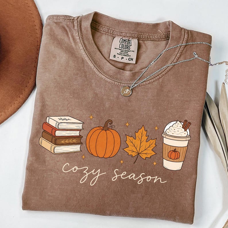 May include: A brown short-sleeved t-shirt featuring a graphic print of a stack of four books, an orange pumpkin, a yellow maple leaf, and a coffee cup with whipped cream. The text "cozy season" is printed in white cursive below the graphics.