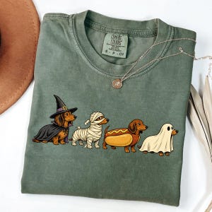 Peut inclure: Un t-shirt &agrave; manches courtes vert olive avec un motif repr&eacute;sentant quatre teckels d&eacute;guis&eacute;s pour Halloween : une sorci&egrave;re, une momie, un hot-dog et un fant&ocirc;me. Le t-shirt pr&eacute;sente une &eacute;tiquette Comfort Colors au col rond int&eacute;rieur.