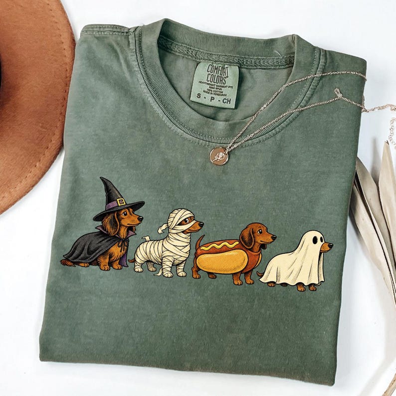 Peut inclure: Un t-shirt &agrave; manches courtes vert olive avec un motif repr&eacute;sentant quatre teckels d&eacute;guis&eacute;s pour Halloween : une sorci&egrave;re, une momie, un hot-dog et un fant&ocirc;me. Le t-shirt pr&eacute;sente une &eacute;tiquette Comfort Colors au col rond int&eacute;rieur.
