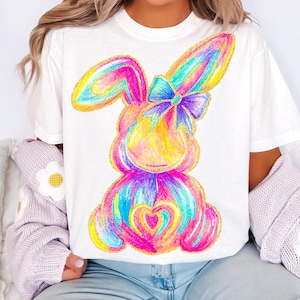 以下が含まれることがあります： 虹色のリボンをつけたカラフルな漫画のウサギが描かれた白いTシャツ。ウサギは虹色で描かれており、胸にハートがあります。