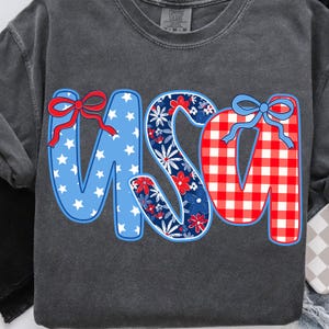 May include: Dark gray sweatshirt with the word "USA" in a patriotic design. The "U" is blue with white stars and a red bow. The "S" is floral with red and blue flowers. The "A" is red and white checkered with a blue bow.