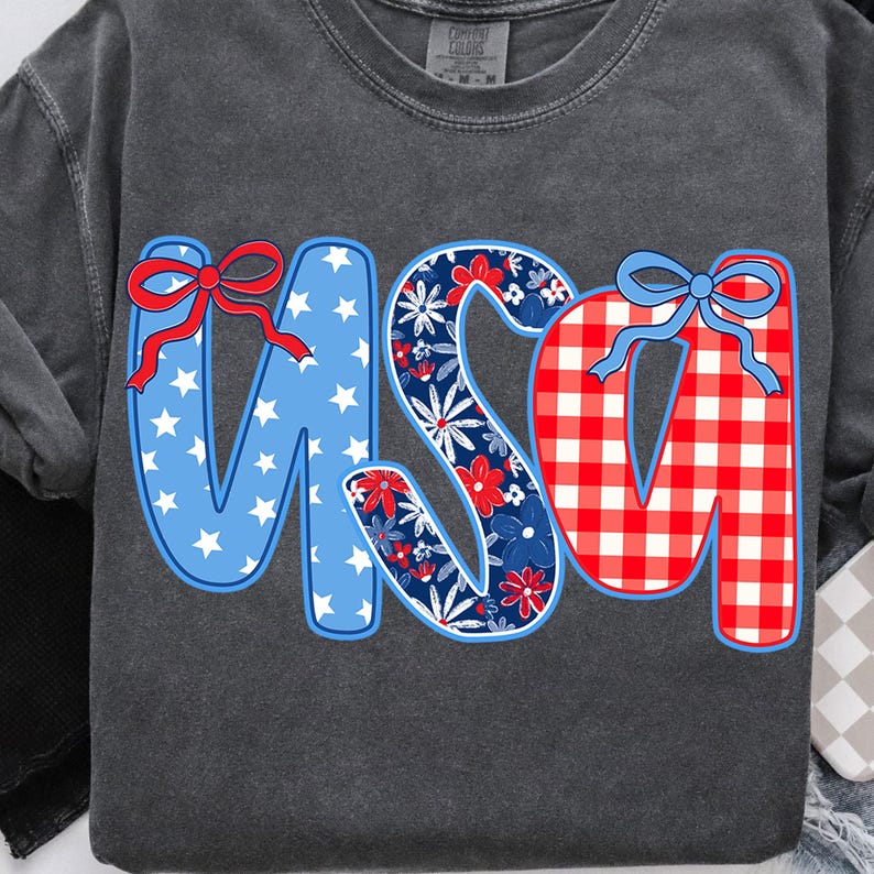 May include: Dark gray sweatshirt with the word "USA" in a patriotic design. The "U" is blue with white stars and a red bow. The "S" is floral with red and blue flowers. The "A" is red and white checkered with a blue bow.