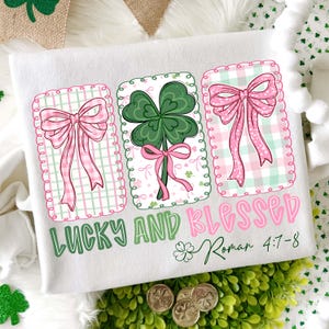 Könnte beinhalten: Weißes T-Shirt mit St. Patrick's Day-Motiv. Es zeigt drei gerahmte Designs: zwei rosa Schleifen und ein grünes Kleeblatt mit einer rosa Schleife. Die Worte "LUCKY AND BLESSED" sind in Grün und Rosa, darunter "Roman 4:7-8".