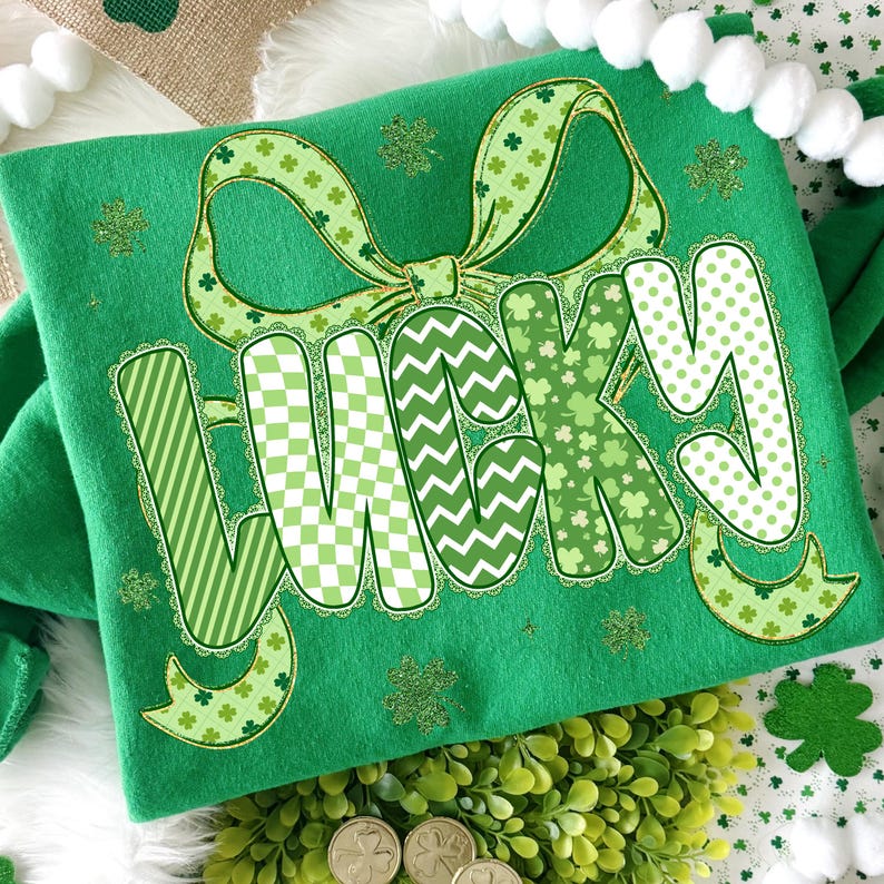 Puede incluir: Una camiseta verde con la palabra "LUCKY" en una variedad de patrones verdes, incluyendo rayas, cuadros y tr&eacute;boles. Un gran lazo con acentos de tr&eacute;bol se encuentra encima de la palabra. La camiseta est&aacute; decorada con purpurina dorada y acentos de tr&eacute;bol.
