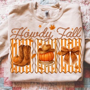 May include: Beige sweatshirt with the text "Howdy Fall" in rope-style lettering. The design includes a pumpkin, cowboy boots, a cowboy hat, and a bow, all in fall colors. The design is printed on a beige sweatshirt.